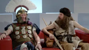 Een foto van een Romeinse veldheer en een man uit de oertijd die samen op een bank zitten. De prehistorische man kijkt naar de Romeinse veldheer. De Romein lacht. 