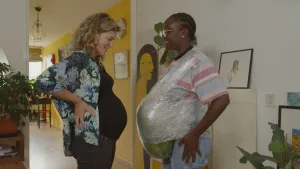 Een foto van presentator Tirsa die met een namaak zwangere buik tegenover een vrouw staat die echt zwanger is en een grote buik heeft.