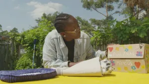 Een foto van presentator Tirsa die aan tafel zit en naar een doos kijkt met de tekst "Rust zacht Minous" erop. De doos heeft ook getekende hartjes, bloemetjes en een vlinder. 