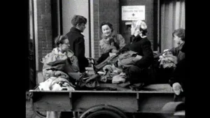 Een zwart-witfoto van vijf mensen bij een kar met kleding en andere stoffen. Ze pakken spullen van de kar en zijn met elkaar in gesprek. De kar is een platte kar op fietswielen. 