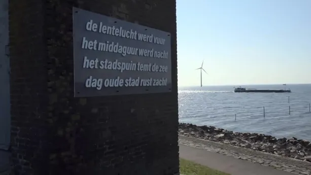 Een foto met links in beeld een stenen muur met daarop een bord met een tekst. Rechts in beeld zie je een weg met daarachter een groot water. In het water staat een windmolen en er vaart een schip voorbij.