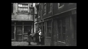 Een foto uit het jaar 1900 van een straat in een stad. Je ziet slecht onderhouden huizen. Op straat staan zes mensen. Het is een somber beeld.