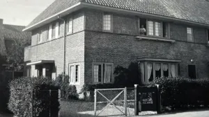 Een zwart-wit foto van een hoekhuis met een tuin en een oprijlaan.  Op de eerste verdieping staat er een man achter een geopend raam.