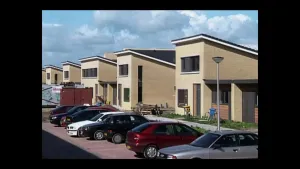 Een foto van een straat met nieuwe huizen. Ze zien er allemaal hetzelfde uit. Voor die huizen zijn parkeerplaatsen met auto's. 