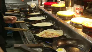 Een foto van 6 pannen met pannenkoeken op een lang fornuis. Er staan een paar mensen bij van wie alleen een stukje voorkant en handen te zien zijn. Achter de pannen staan stapels met borden.