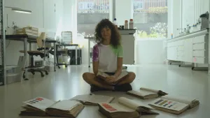 Een foto van presentator Serah. Ze zit in een kantoor op de grond. Voor haar liggen een paar opengeslagen boeken. Daarin zie je allemaal verschillende kleuren. Ze heeft ook een open boek op schoot. 