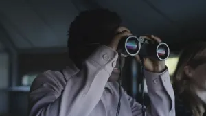 Een foto van een man die in een gebouw staat en met een verrekijker naar buiten kijkt. Er staat nog iemand naast hem die ook naar buiten kijkt.