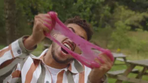 Een foto van presentator Nizar. Hij heeft een hoeveelheid roze slijm vast in zijn handen. Hij trekt het slijm uit elkaar en schreeuwt daarbij.