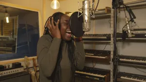 Een foto van presentator Tirsa die in een microfoon zingt in een muziekstudio. Op de achtergrond zijn er verschillende digitale piano's te zien.