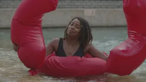 Een foto van presentator Eva. Ze staat in het water, kijkt in de camera, en houdt twee grote rode ballonnen vast. De ballonnen hebben allebei de vorm van een maansikkel. Zelf heeft ze een rode ronde zwemband om zich heen.