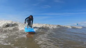 Een foto van presentator Eva op een surfplank in de zee. Ze surft naar de camera toe. Ze zit met een knie op de surfplank, en leunt met een hand op de surfplank. Achter haar zie je een golf. 
