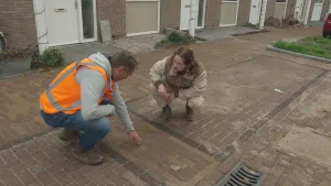 Een foto van de presentator en een stratenmaker. Ze zitten gehurkt op straat. De stratenmaker wijst naar de straatstenen in de weg. De presentator kijkt naar de stratenmaker.