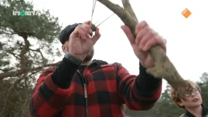 Een foto van boswachter Tim die met een katapult een dennenappel weg wil schieten. Een jongetje kijkt mee met hem. 