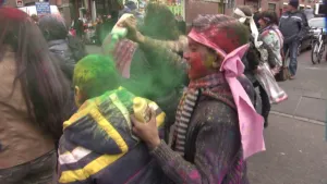 Een foto van mensen die Holi vieren op straat. Ze zitten onder het poeder van verschillende kleuren. De jongen rechts vooraan op de foto heeft een roze lapje stof om zijn hoofd en strooit groen poeder uit een flesje over het hoofd van de voorste jongen.