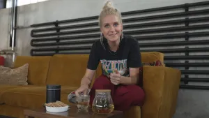 Een foto van de presentator. Ze zit op een bank en kijkt in de camera. Voor haar staan op een tafeltje een theepot, een glas thee en een bakje met koekjes. 