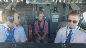 Een foto van de binnenkant van de cockpit van een vliegtuig. Op de voorgrond zie je twee piloten zitten met koptelefoons op. Op de achtergrond zit de presentator. Zij en een van de piloten kijken elkaar aan.