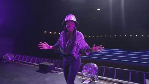 Een foto van presentator Tirsa die voor de camera staat op een podium. Er schijnt paars licht op haar en het podium.