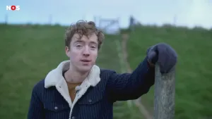 Een foto van een presentator die op een stenen trap in het midden van een groene heuvel met gras staat. Hij draagt een blauwe jas met een witte kraag. Zijn blauw gehandschoende linkerhand ligt op een grijze houten paal aan de rechterkant van de foto.