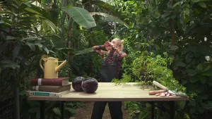 Een foto van de presentator die achter een tafel staat. De presentator heeft haar handen bij haar mond om iets te roepen. De tafel staat tussen allemaal bomen. Op de tafel liggen rode kolen, tuingereedschap, boeken en een gieter.