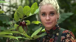 Een foto van presentator Janouk die naast de plant Aristolochia staat. De plant bestaat naast takken en groene bladeren uit een opvallende rode bloem met een holle ingang. De bloem heeft drie lange, rode, en dunne uiteinden die naar beneden kronkelen.