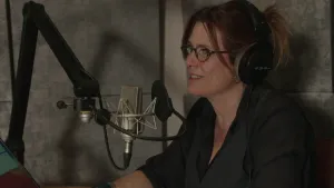 Een foto van een vrouw achter een microfoon. Ze heeft een grote koptelefoon op. Ze zit vlak bij een muur met grote grijze platen erop.