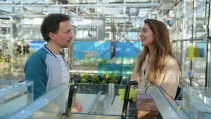 Een foto van een presentator die met onderzoeker Fee Smulders in een plantenkas van glas staat. Op de achtergrond staan kleine groene planten in de aarde in een bak. Op de voorgrond zijn de bovenkanten van plastic bakken te zien en twee rode bakstenen. 