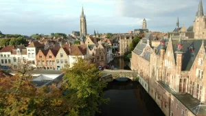 Een foto van de stad Brugge in België. Naast een gracht met water en een brug staan rechts oude gebouwen. Eén gebouw heeft twee torens met grijze daken en ronde, roodkleurige openingen eronder. Op de foto staan ook huizen en drie torens in de achtergrond.