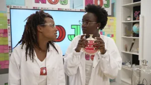 Een foto van presentatoren Eva en Tirsa die een witte laboratoriumjas aanhebben over hun "NOJ" t-shirt. Ze hebben ook een doorzichtige veiligheidsbril op. Tirsa staat rechts op de foto en heeft in haar handen een scheetkussen vast. 