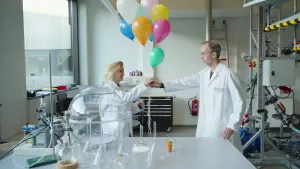 Een foto van presentator Janouk en Marcel die in een laboratorium staan. Marcel heeft een tros ballonnen van allerlei kleuren gevuld met helium vast in zijn hand. Janouk pakt de ballonnen over op de foto. Ze dragen allebei een witte laboratoriumjas.