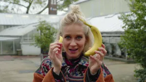 Een foto van presentator Janouk die een bananensnoepje in haar rechterhand omhoog houdt en een 'echte' banaan van de fruitsoort in haar linkerhand omhoog houdt voor de camera. Janouk kijkt recht in de lens terwijl ze spreekt.