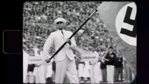 Een zwart-wit foto van een man in wit pak en witte pet die met de vlag van Nazi Duitsland zwaait in een stadion. Op de tribunes zit allemaal publiek en achter de man staan meer mensen. De foto is genomen tijdens de Olympische Spelen van 1936 in Berlijn.