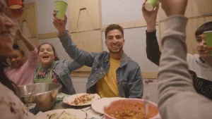 Een foto van de presentator met een paar kinderen. Ze zitten aan een tafel met eten. Ze proosten allemaal. Ze houden hun drinkbekers feestelijk omhoog. 