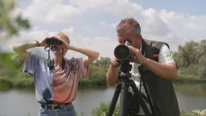 Een foto van twee mensen die door verrekijkers kijken. En van de verrekijkers is heel groot en staat op een standaard. Op de achtergrond is natuur te zien met een meer.