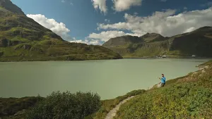 Een foto van presentator Nizar die aan de rand van een groot stuwmeer staat. Hij is iets aan het uitleggen. Op de achtergrond zie je bergen en een blauwe lucht met wolken.