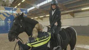 Een foto van de presentator op een paard. Ze kijkt wat angstig. Op de voorgrond zie je een politieagent met haar arm richting de presentator. Op de achtergrond zie je een groot doek met daarop het woord "POLITIE".