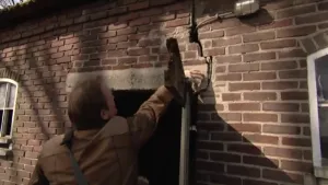 Een foto van de buitenkant van een muur van een oud huis. In die muur zit een grote scheur boven een deur. Een man houdt zijn hand vlakbij die scheur. 