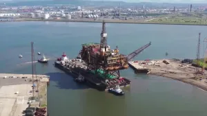 Een foto van een boorplatform gezien vanuit grote hoogte. Het boorplatform staat op een groot drijvend voorwerp in een haven. Eromheen zie je sleepboten die het boorplatform verplaatsen.
