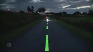 Een foto van een fietspad in het donker. Je staat op het fietspad. De onderbroken strepen in het midden lijken lichtgevend groen te zijn. Een stukje verderop staat een man gebukt. Hij schijnt met een felle lamp op een van die strepen van het fietspad.
