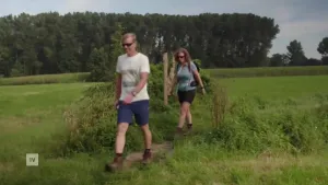 Een foto van een man die in de natuur op een klein paadje loopt. Het is mooi weer. Achter hem loopt een vrouw met een rugzak. Ze lopen tussen de weilanden met op de achtergrond bomen. 