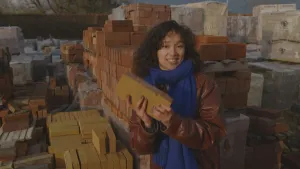 Een foto van presentator Serah. Ze houdt een baksteen in haar handen en kijkt in de camera. Op de achtergrond zie je hele stapels van bakstenen. Het zijn wel duizenden bakstenen. 