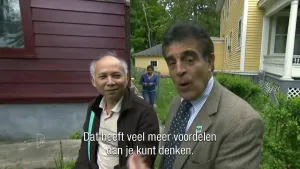Een foto van twee mannen die in de camera kijken. Een man is wat aan het vertellen. Hij heeft een jasje met stropdas aan. Op de achtergrond zie je enkele houten huizen. Daartussen loopt een vrouw. Verder zie je bomen, struiken en gras.