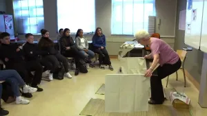 Een foto van Thea Meulders in een klaslokaal. Links in beeld zitten scholieren. Ze kijken naar haar. Thea staat gebogen en heeft twee witte doeken vast.  Daaronder liggen bamboematten.