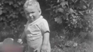 Een foto in zwartwit van een peuter die buiten staat en een stoelleuning vasthoudt. Hij kijkt je aan. Op de achtergrond zie je struiken.