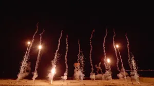 Een foto van een vuurwerkshow in het donker waarbij negen stukken vuurwerk omhoogschieten of al omhooggeschoten zijn. Bij vijf stukken vuurwerk die omhoogschieten zijn heldere geel-oranje lichtjes zichtbaar, bij de vier die al opgestegen zijn rookwolken.