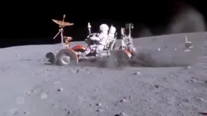 Een foto van een maanwagen die op de maan rijdt. Er zit een astronaut op die de wagen bestuurt. Achter de wielen stuift het maanzand meters omhoog. De hemel is zwart van kleur. 