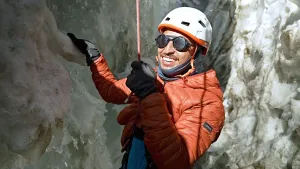 Een foto van presentator Nizar die aan een klimtouw hangt, en glimlacht. Hij heeft handschoenen en een oranje jas aan en een witte helm op. Hij hangt in een gletsjerspleet.