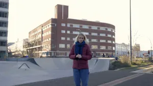 Een foto van presentator Janouk. Ze staat op straat en vertelt iets aan de kijker. Op de achtergrond zie je een skate-baan en een paar gebouwen.