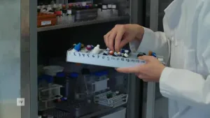 Een foto van een arts in een witte jas. Hij heeft een bakje vast met daarin allerlei kleine flesjes. Hij heeft een van die flesjes vast. Op de achtergrond zie je een laboratoriumkast met daarin nog veel meer flesjes.