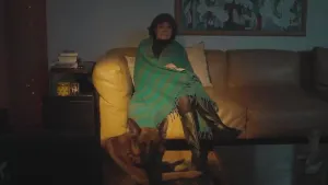 Een foto van een donkere kamer. Op de bank zit een vrouw met een deken om haar heen. Op de grond zit een hond. 