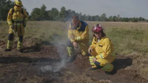 Een foto van drie brandweermannen in gele uniformen. Ze zijn in een natuurgebied. Voor hen zie je een stuk verbrandde aarde waar rookt uit komt. 